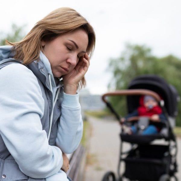 mulher sentada no branco triste, com carrinho de bebê ao fundo, representando sentimento de tristeza do baby blues