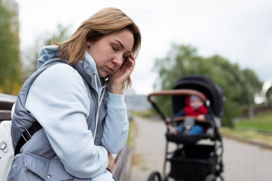 mulher sentada no branco triste, com carrinho de bebê ao fundo, representando sentimento de tristeza do baby blues